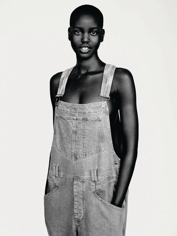 Portrait of a stylish young Black woman in denim overalls. The image is a close-up shot in black and white with soft studio lighting.