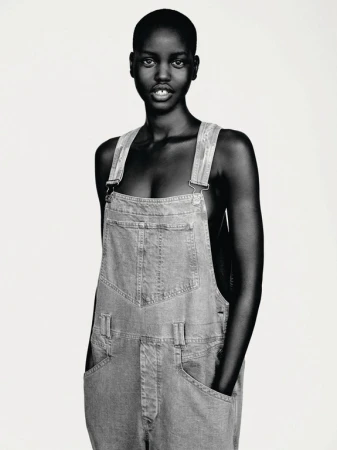 Portrait of a stylish young Black woman in denim overalls. The image is a close-up shot in black and white with soft studio lighting.
