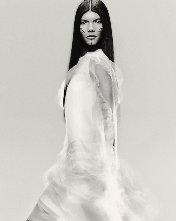 Fashion portrait of a woman with face makeup and a sheer white garment in a studio with soft lighting and a neutral backdrop.