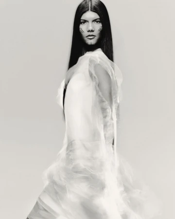 Fashion portrait of a woman with face makeup and a sheer white garment in a studio with soft lighting and a neutral backdrop.