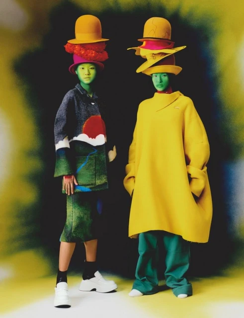 Two people in colorful hats and outfits stand side-by-side with green tinted skin, striking a pose against a dark background with yellow highlights.