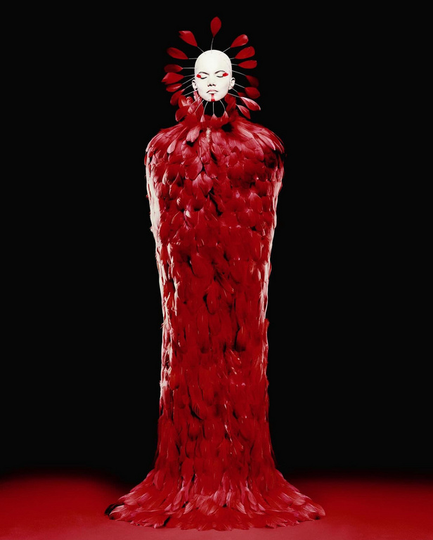 Figure in a long, red, feathered gown, white face and headdress with red accents, standing in a studio with a dark background.