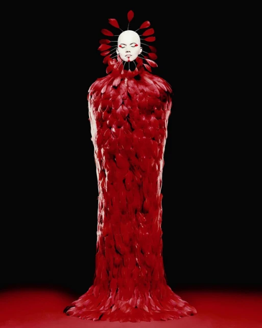 Figure in a long, red, feathered gown, white face and headdress with red accents, standing in a studio with a dark background.