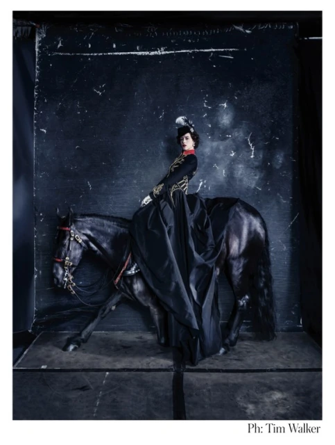 A woman in vintage attire rides a black horse against a dark backdrop in a fashion editorial. The horse is in motion, captured mid-stride.