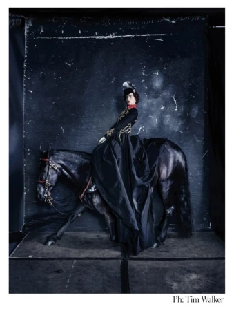 A woman in vintage attire rides a black horse against a dark backdrop in a fashion editorial. The horse is in motion, captured mid-stride.