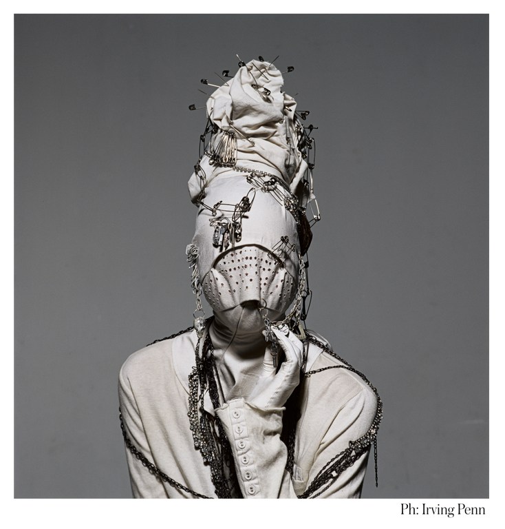 Abstract fashion portrait of an anonymous person in a white outfit, head wrapped in fabric and decorated with safety pins and chains, studio setting.