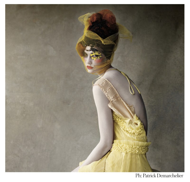 Woman in yellow couture dress, headpiece, and makeup sits in front of a gray background in a fashion photography studio shot.