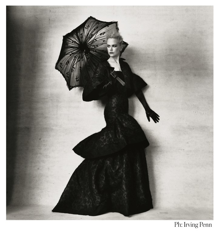 A woman in a black gown and gloves stands against a textured backdrop, holding a black lace parasol, evoking a vintage editorial style.