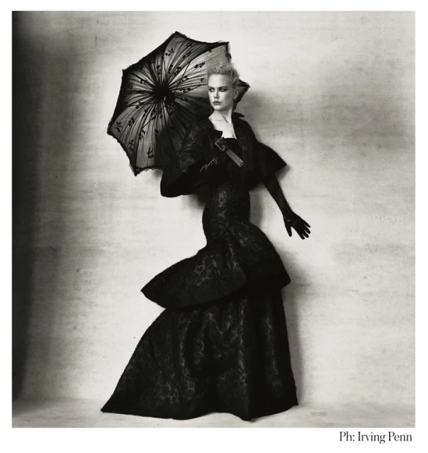 A woman in a black gown and gloves stands against a textured backdrop, holding a black lace parasol, evoking a vintage editorial style.