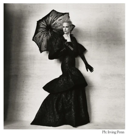 A woman in a black gown and gloves stands against a textured backdrop, holding a black lace parasol, evoking a vintage editorial style.