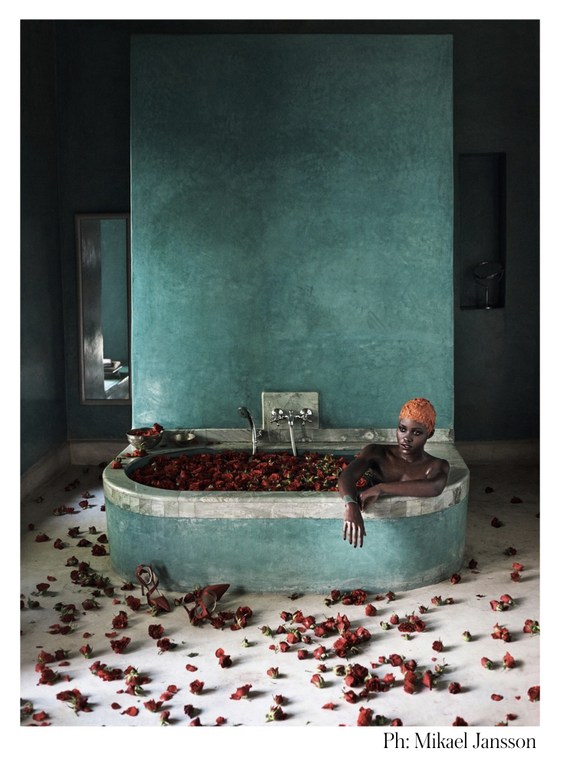 Black woman in a turquoise bathtub filled with rose petals, in a dark-toned bathroom with rose petals scattered on the floor.