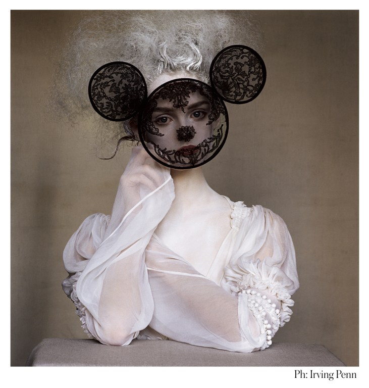 Conceptual fashion shot features an adult woman with pale skin wearing a sheer white gown and a lacy Mickey Mouse mask, in a studio setting.