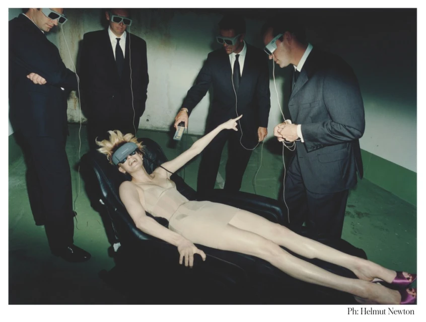 Futuristic fashion photography featuring a woman lying on a chair surrounded by men in suits and VR headsets in a stark setting.