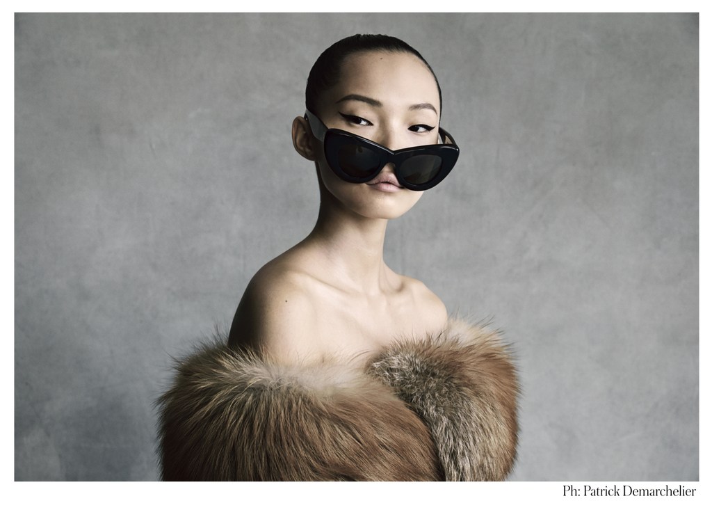 Portrait of an Asian woman with a shaved head wearing oversized black sunglasses and a brown fur shawl against a gray backdrop.