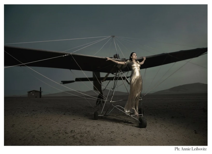 Fashion shot of a woman in a gold gown standing on an airplane wing in a desert landscape against a stormy sky. By Annie Leibovitz.
