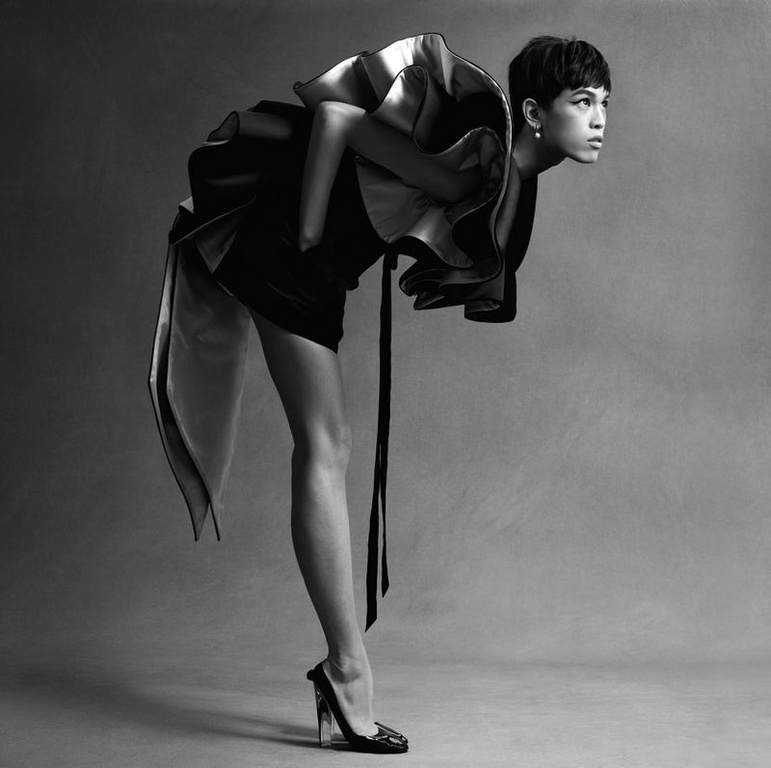 Monochrome fashion shot of a slender adult with short dark hair wearing a dramatic ruffled dress, leaning forward with a high-heeled shoe.