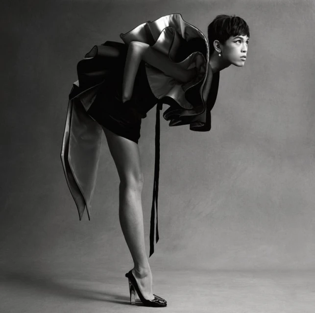 Monochrome fashion shot of a slender adult with short dark hair wearing a dramatic ruffled dress, leaning forward with a high-heeled shoe.