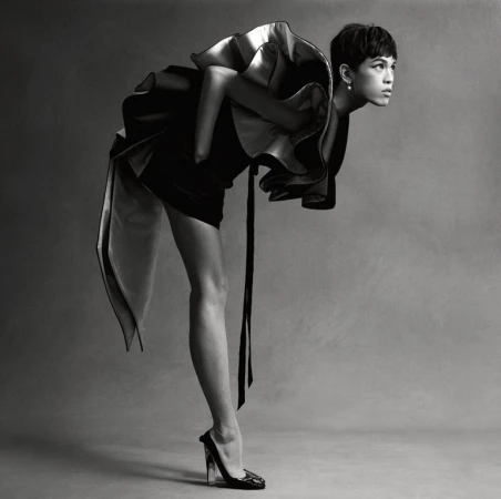 Monochrome fashion shot of a slender adult with short dark hair wearing a dramatic ruffled dress, leaning forward with a high-heeled shoe.