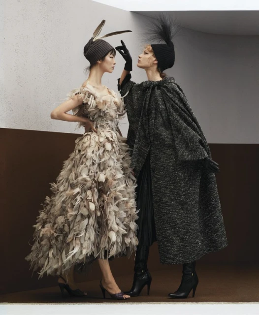 Two fashion models in a photo studio. One model wears a feathered dress and the other wears a gray coat and a black beanie.