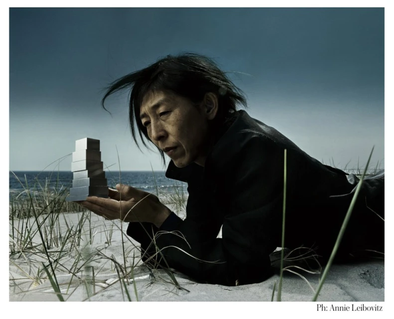 Woman lying in the sand with a tower of mirrored blocks, the ocean is in the background under a cloudy sky.