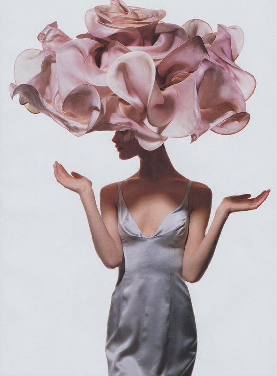 Woman in a metallic silver slip dress wearing an oversized ruffled pink floral headpiece, posed against a white background.