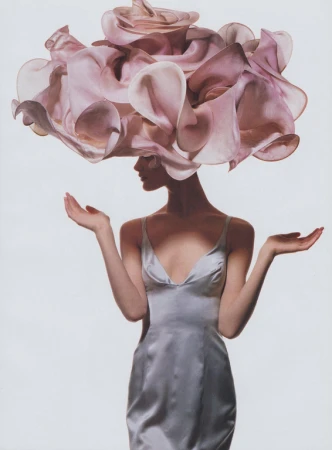 Woman in a metallic silver slip dress wearing an oversized ruffled pink floral headpiece, posed against a white background.