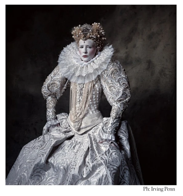 Portrait of a woman wearing a large Elizabethan era white gown, pearls, and a crown against a dark background in a photo studio.