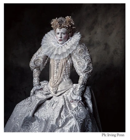Portrait of a woman wearing a large Elizabethan era white gown, pearls, and a crown against a dark background in a photo studio.