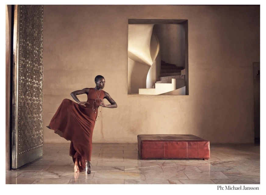 Woman in flowing dress strikes a pose in a building interior with a spiral staircase visible through a window, bathed in warm, natural light.