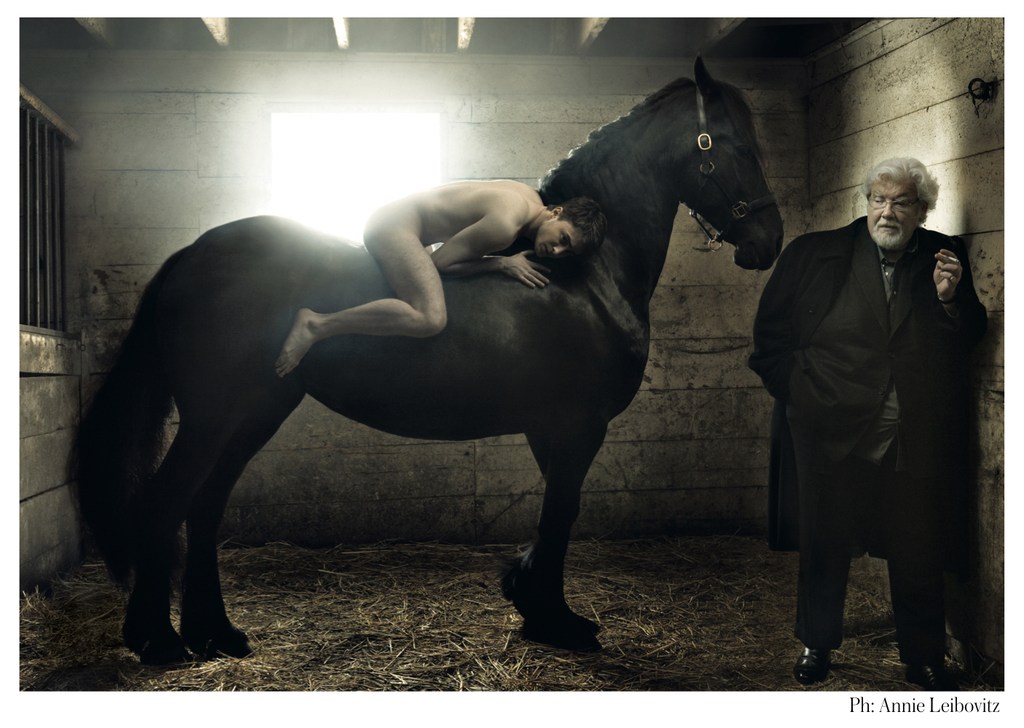 Striking portrait of a bare-chested man reclining on a glossy black horse next to an older man in a dark coat inside a stable setting.
