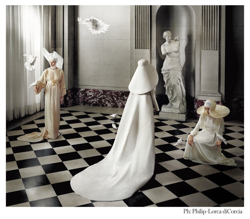 Three women in flowing gowns and large hats stand on a black and white checkered floor with white doves in an opulent indoor setting.