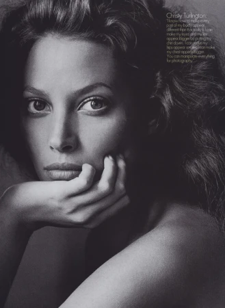 Striking black and white portrait of Christy Turlington with dark wavy hair, her chin resting on her hand in a classic pose.
