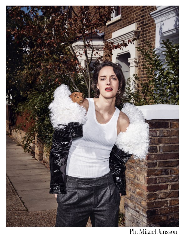 Portrait of Phoebe Waller-Bridge wearing a white tank top, grey trousers and a black and white jacket, posing outside a brick building.