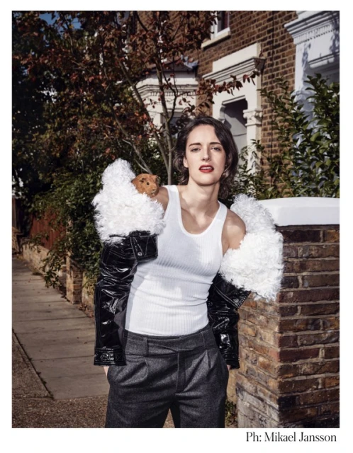 Portrait of Phoebe Waller-Bridge wearing a white tank top, grey trousers and a black and white jacket, posing outside a brick building.