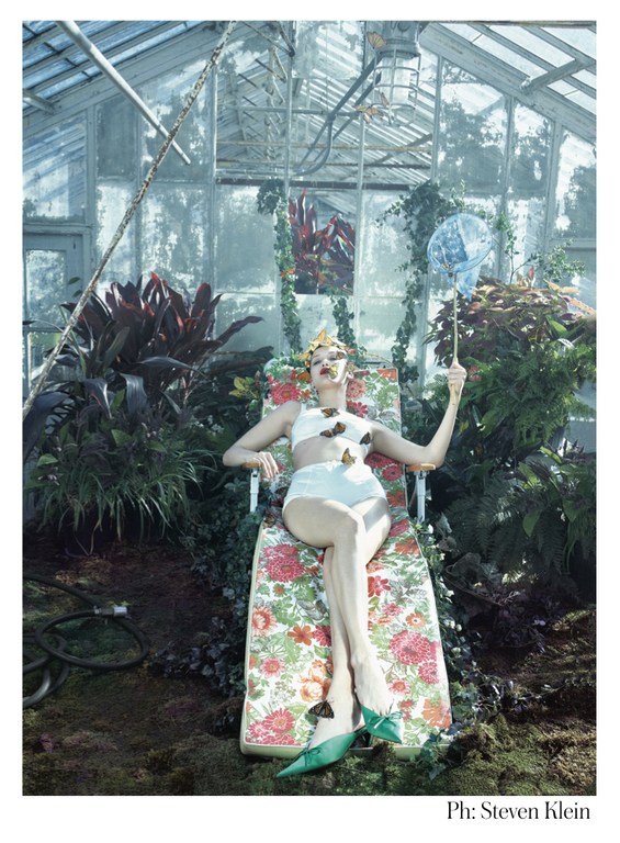 A woman in a vintage swimsuit and green heels lounges in a greenhouse, surrounded by plants and butterflies, holding a butterfly net.