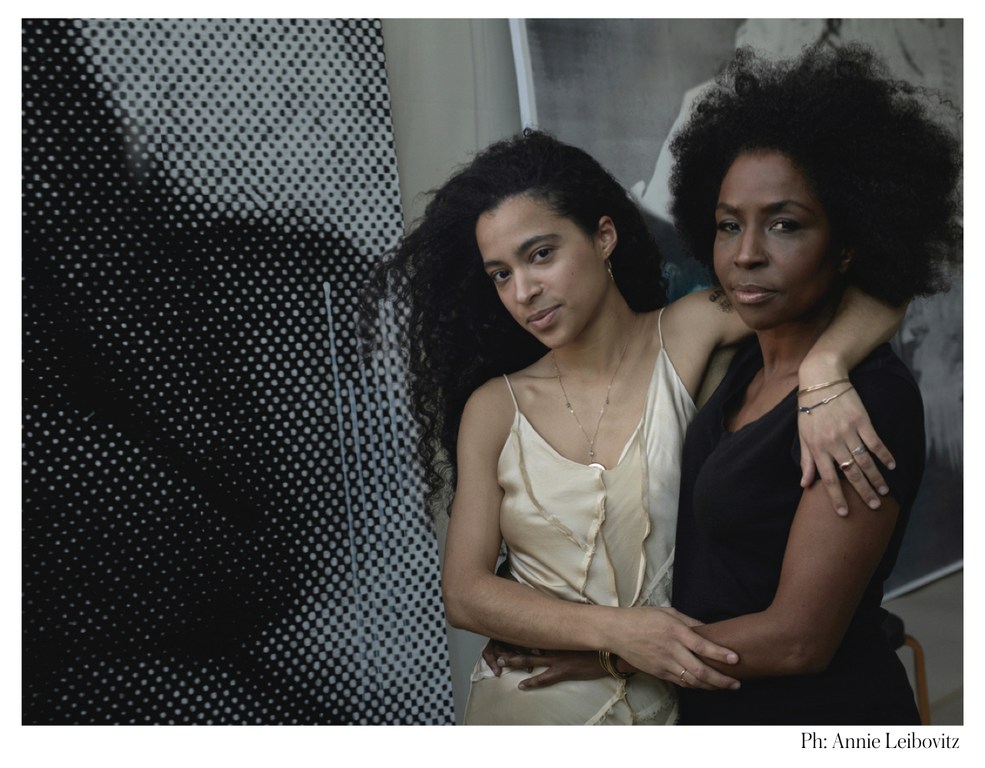 A portrait captures two women with natural hair indoors, one in a cream dress, the other in black, posed against a striking patterned backdrop.