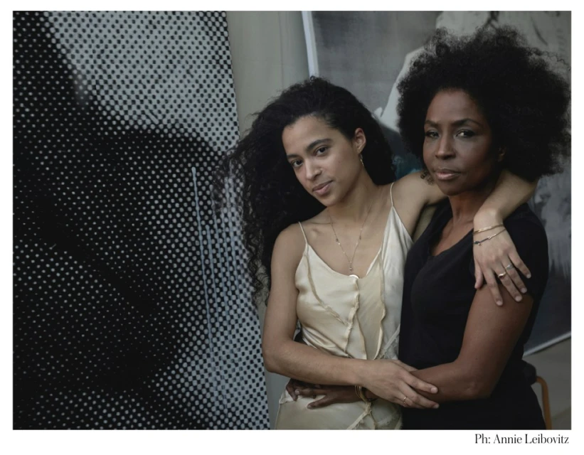 A portrait captures two women with natural hair indoors, one in a cream dress, the other in black, posed against a striking patterned backdrop.