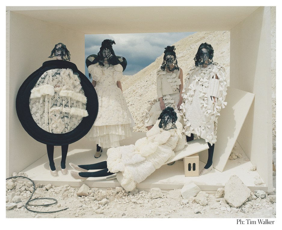 High fashion editorial image featuring five models in conceptual white dresses and lace masks, standing in a minimalist off-white box set against a desert backdrop.