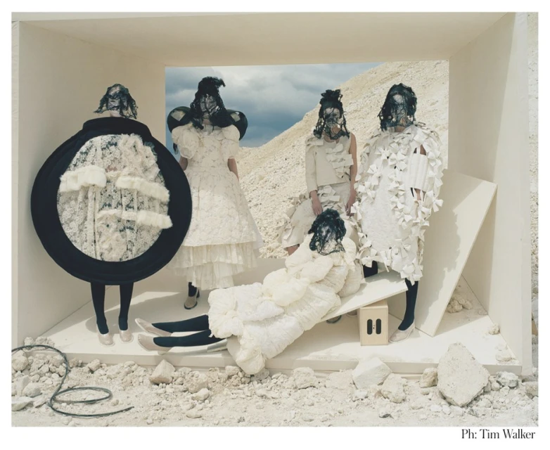 High fashion editorial image featuring five models in conceptual white dresses and lace masks, standing in a minimalist off-white box set against a desert backdrop.