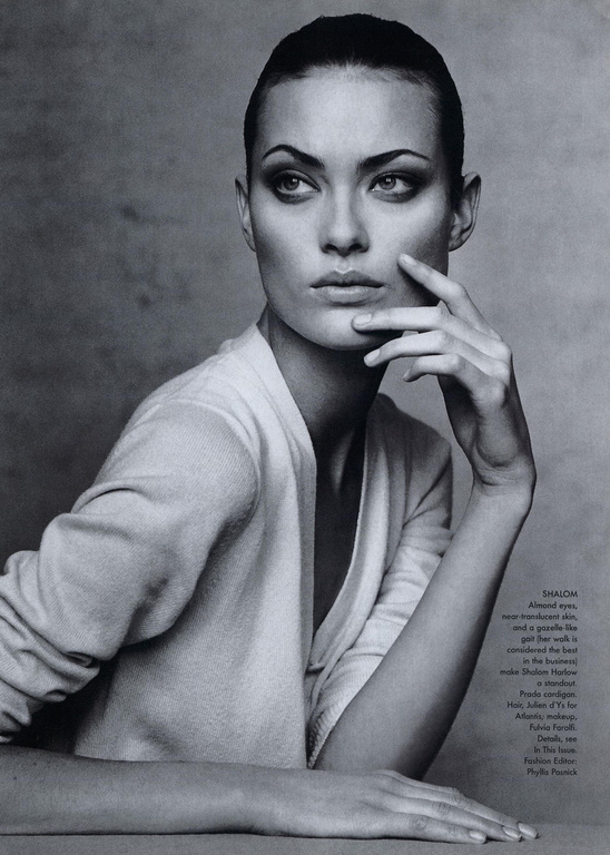 Monochromatic portrait of a woman with slicked-back hair wearing a v-neck top, gazing to the side with her hand near her face.