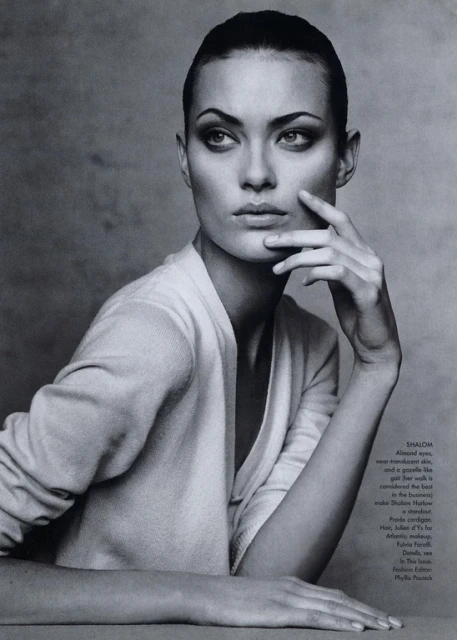 Monochromatic portrait of a woman with slicked-back hair wearing a v-neck top, gazing to the side with her hand near her face.