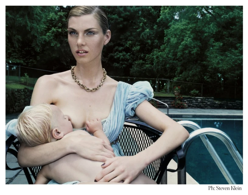 Adult female sitting poolside in a black wire chair breastfeeding a young blond child in an outdoor setting with trees and a stone wall.