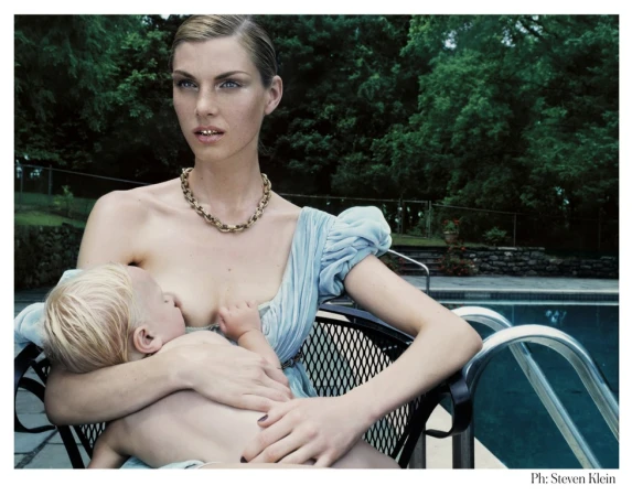 Adult female sitting poolside in a black wire chair breastfeeding a young blond child in an outdoor setting with trees and a stone wall.