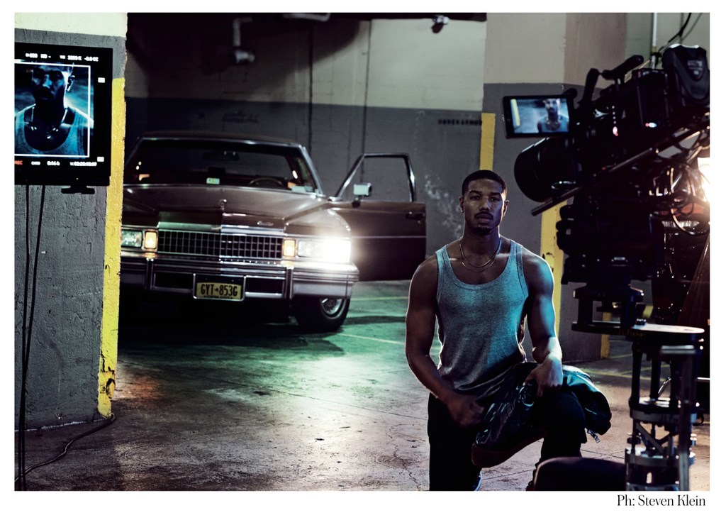 Michael B. Jordan poses next to a vintage car on a film set with studio lighting and film equipment visible, creating a behind-the-scenes ambiance.