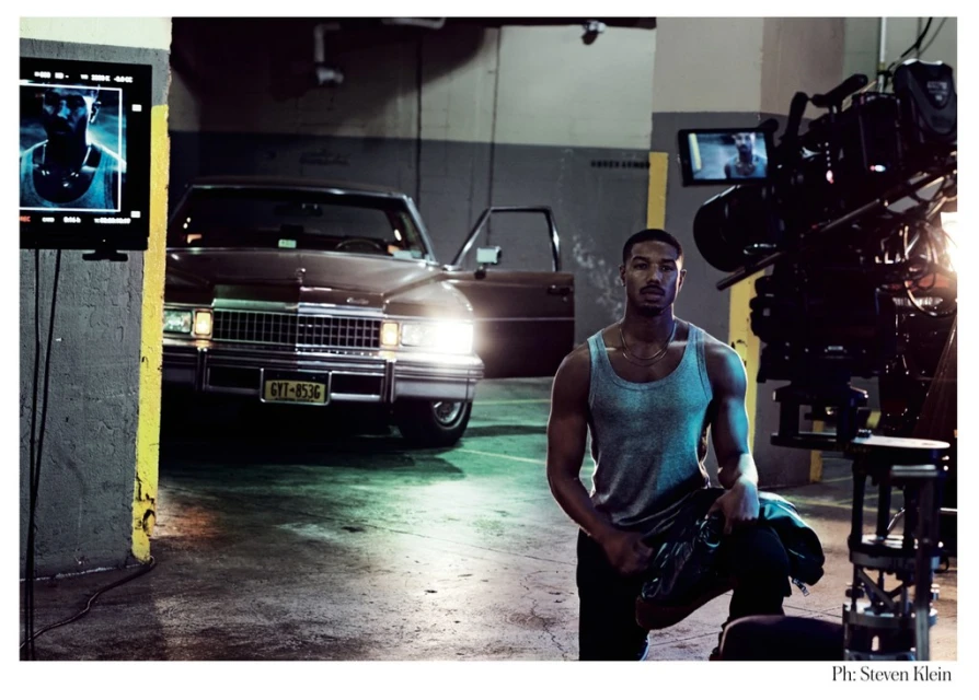 Michael B. Jordan poses next to a vintage car on a film set with studio lighting and film equipment visible, creating a behind-the-scenes ambiance.