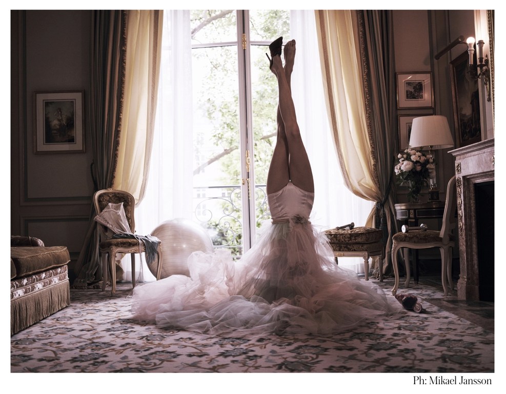Upside-down view of a woman in a pink dress and black high heels performing a handstand in a decorated room near a large window.