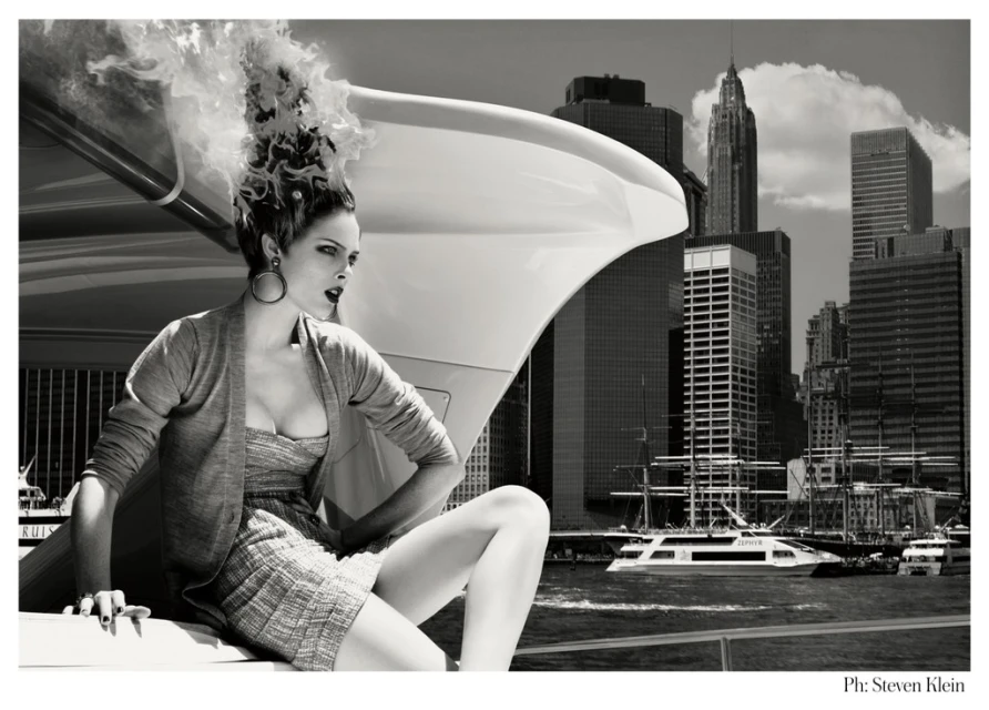 Black and white fashion shot features a woman sitting atop a white vehicle with a cityscape backdrop, creating a dramatic editorial scene.
