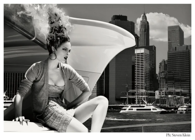 Black and white fashion shot features a woman sitting atop a white vehicle with a cityscape backdrop, creating a dramatic editorial scene.