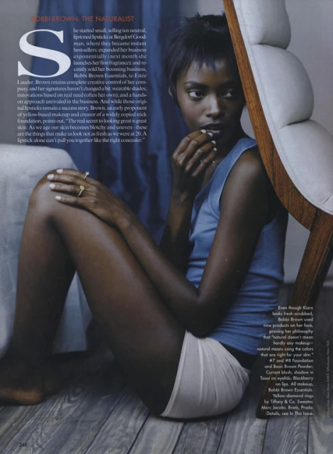 Dark-skinned young woman in studio, seated on floor with arm resting on leg and fingers near lips. Hair short, top blue, and chair visible.