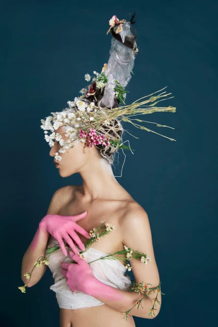 Woman with flowers in hair and pink gloves poses in front of a dark blue backdrop in this artistic beauty fashion shot.